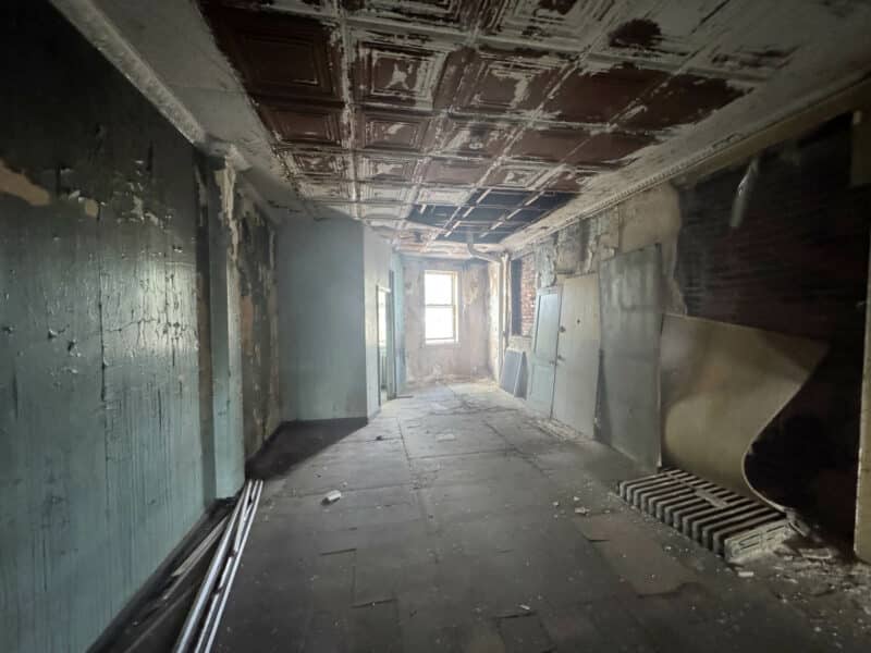 Narrow upper-floor corridor with distressed ceiling and peeling paint, illuminated by natural light.