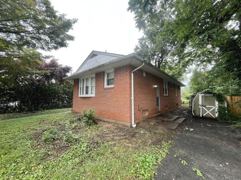 Side view of brick bungalow at 5900 Winthrope Avenue in Baltimore, with asphalt driveway, shed, and surrounding mature trees.