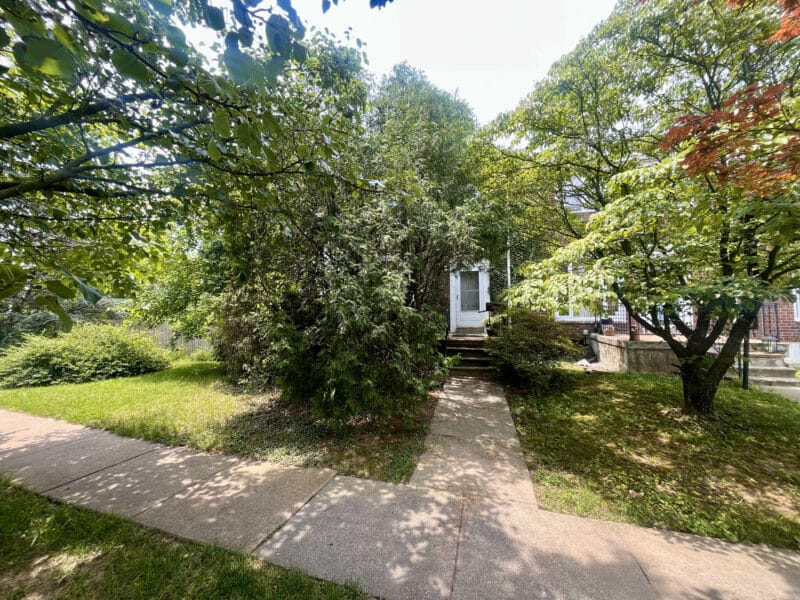 Wider angle front view of brick townhome in Woodlawn, MD, with concrete walkway, overgrown trees, and front porch partially visible.