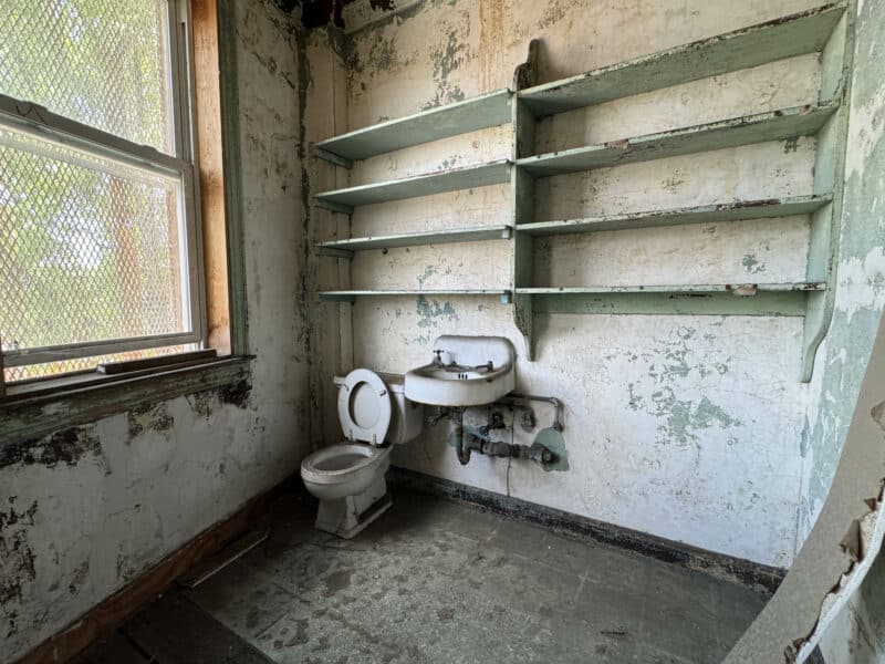 Upper-floor bathroom with pedestal sink, toilet, and open shelving in distressed condition.