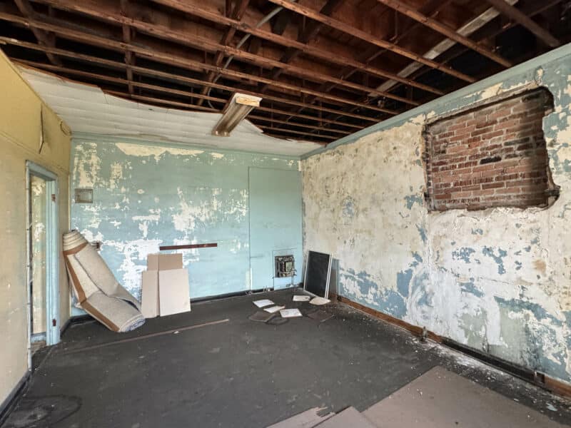 Vacant interior room with exposed brick, peeling paint, and partial ceiling removal.