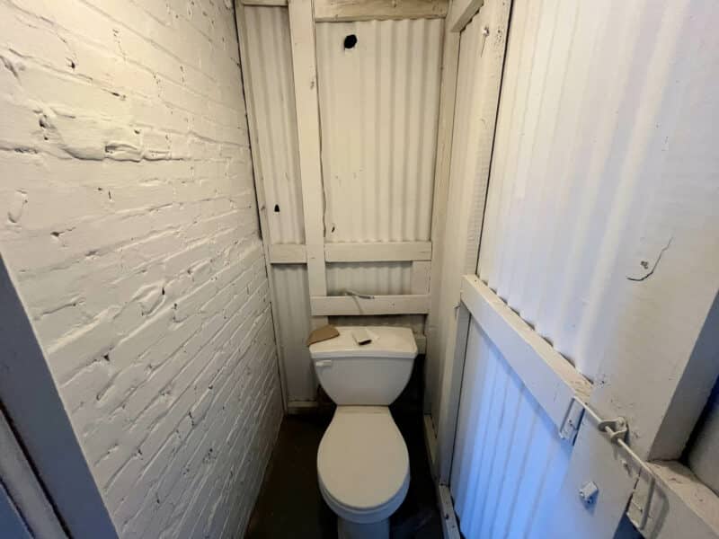 Toilet stall in basement with painted brick and wood panel walls at 5839 Belair Road.