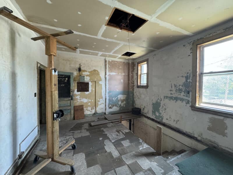 Room with partially finished ceiling, peeling paint, large windows, and a stairwell leading down.