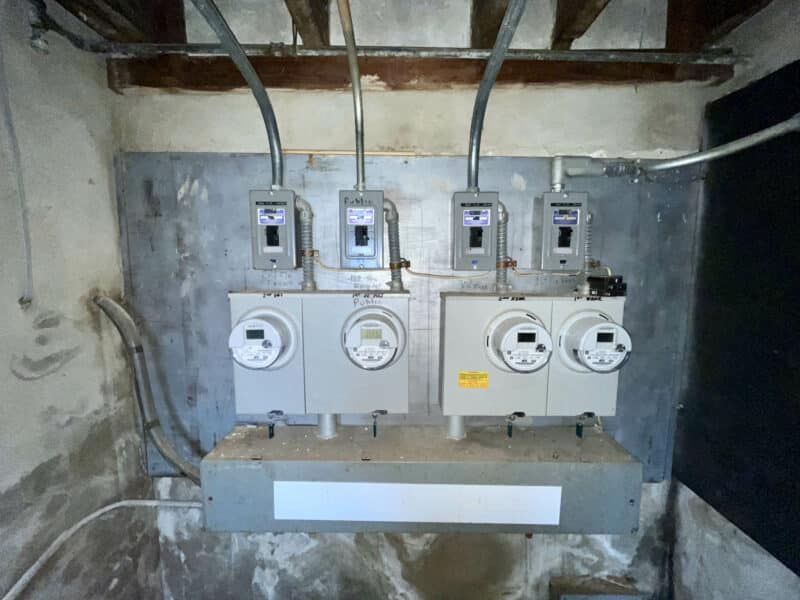 Electric meter bank with four meters and service panels mounted on concrete wall in the basement at 5839 Belair Road.