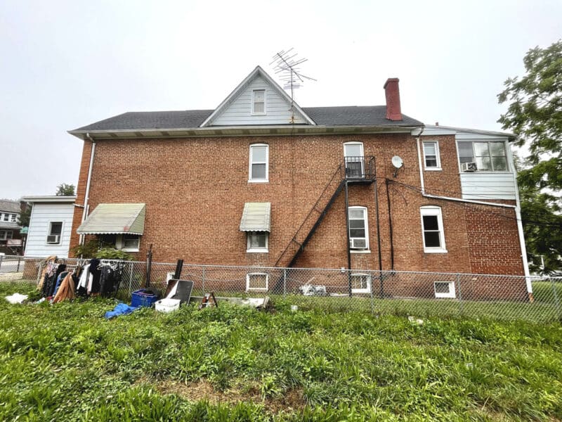 Side view of 5839 Belair Road showing multi-level brick structure with fire escape, fenced yard, and window-mounted AC units.
