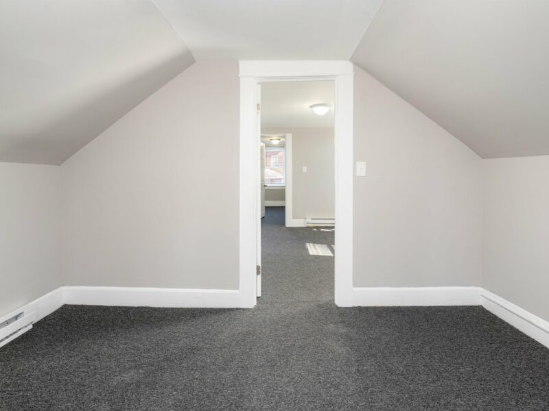 Upstairs bedroom with fresh carpet, sloped ceilings, and a view into adjacent rooms in a renovated Baltimore bungalow.