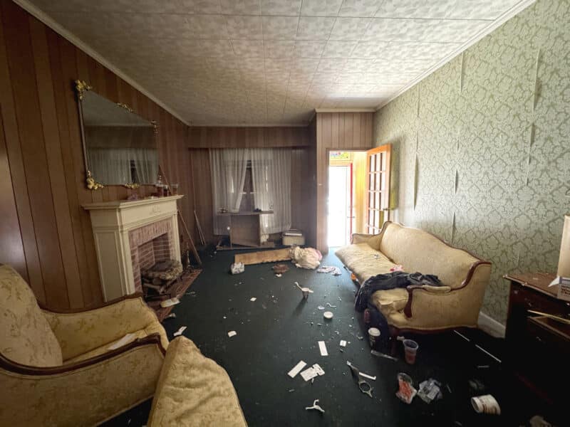 Interior view of the living room with vintage furnishings, decorative fireplace, wall paneling, and scattered debris in a Southeast Baltimore rowhome.