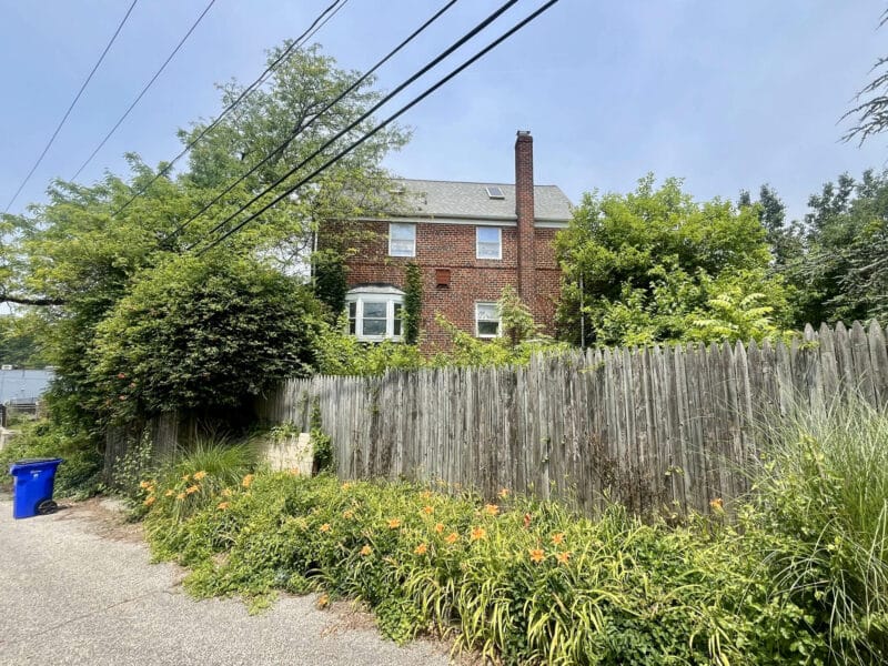 Side view of brick end-of-group townhome with overgrown fence line and alley access in Woodlawn, Baltimore County.