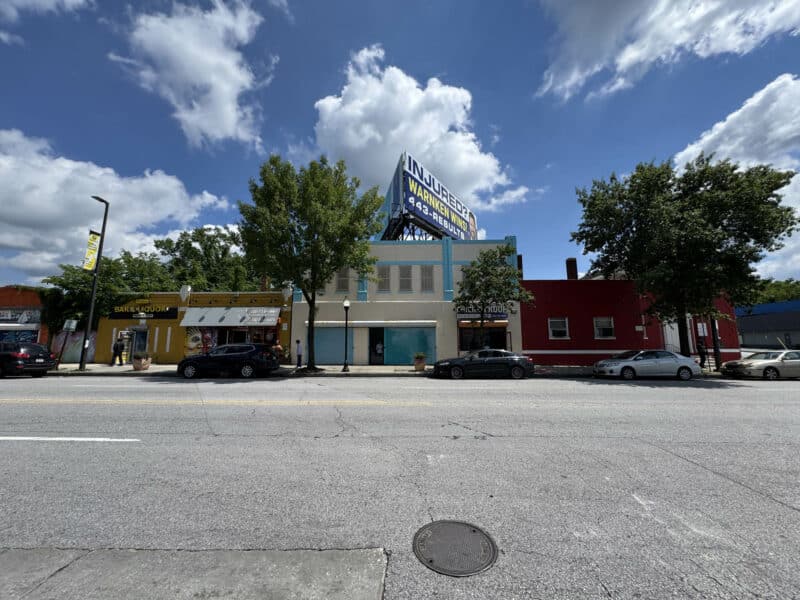 Wide-angle view of commercial building at 5109 Park Heights Avenue with billboard, adjacent storefronts, and cars parked along busy street in Northwest Baltimore.