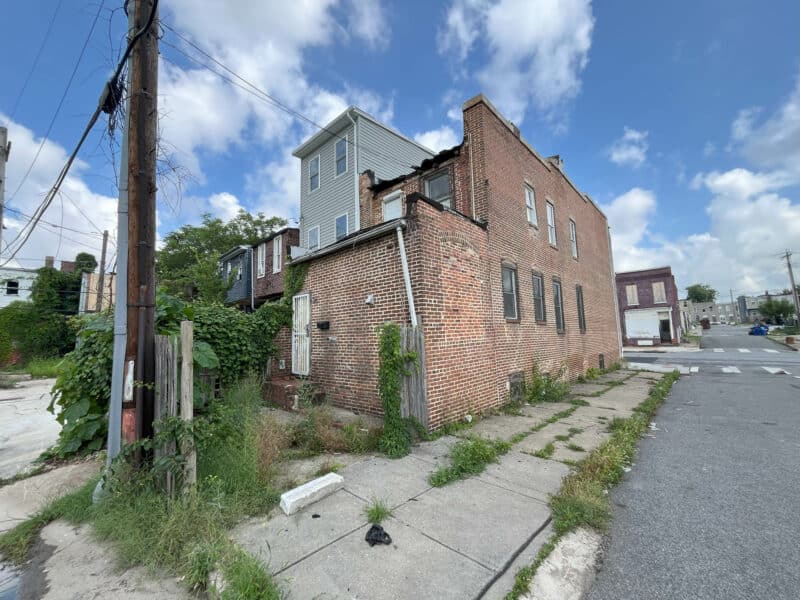 Rear and side view of 2307 E. Preston Street in Broadway East, showing overgrown vegetation and brick exterior with visible deterioration.