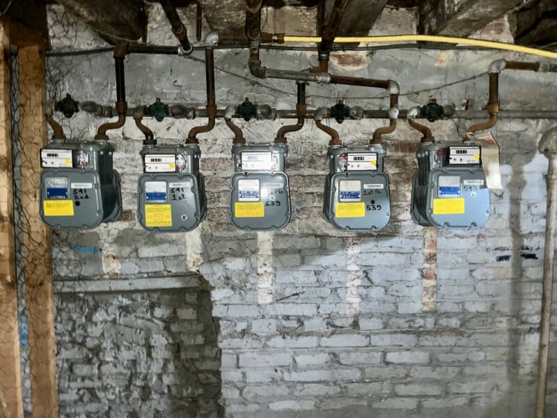 Five gas meters mounted on exposed brick basement wall with overhead piping