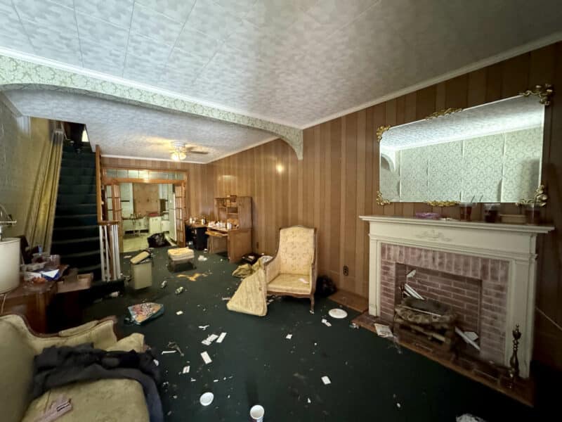 Wide-angle interior photo showing living and dining rooms, decorative ceiling, wood-panel walls, brick fireplace with mirror, green carpet, stairway, and debris in a Highlandtown area townhome.