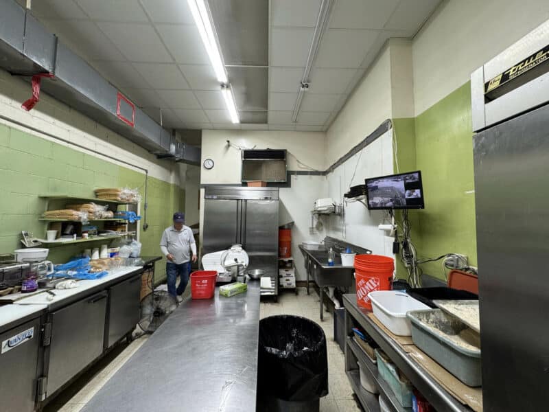 Interior view of a commercial kitchen with stainless steel prep stations, open shelving, and active food preparation.