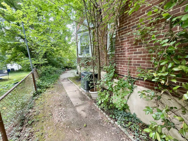 Side walkway along a brick end-unit townhome with overgrown vegetation and a chain-link fence.