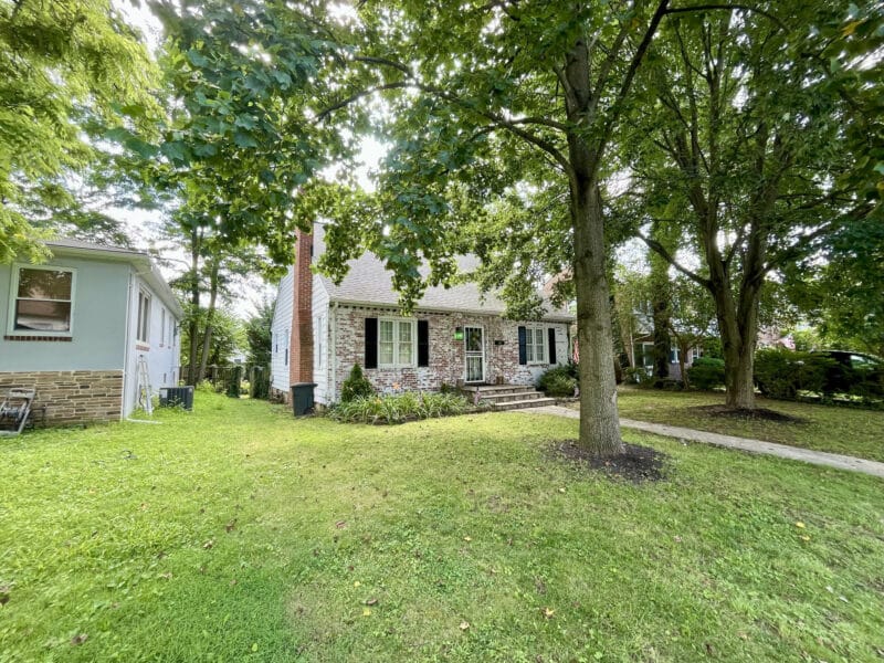 Side yard and partial front view of a brick and siding bungalow with mature trees and landscaped lawn.