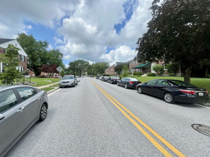Residential street lined with parked cars and red brick townhomes under a partly cloudy sky.