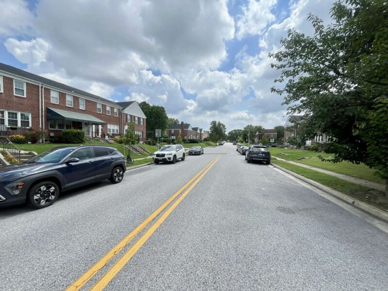 View down a suburban residential street lined with brick townhomes, green lawns, and parked cars under a cloudy sky.