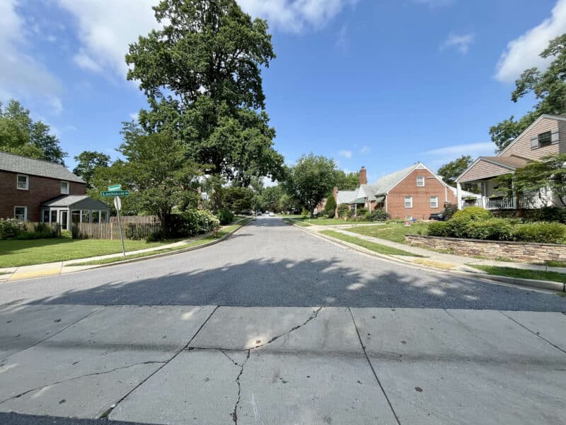 Street view of Lochinvar Drive in Catonsville, showing tree-lined sidewalks, single-family brick homes, and a blue sky with scattered clouds.