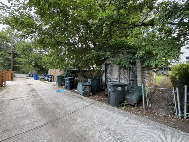 Wide rear alley view showing trash bins, garages, and gated yard access behind townhomes.