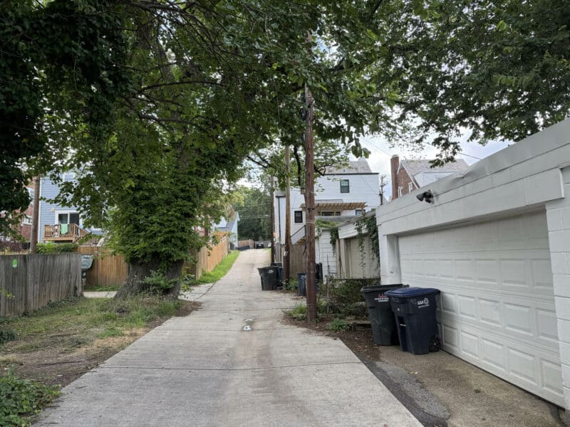Paved rear alley lined with garages, trash bins, and adjacent fences behind residential buildings.
