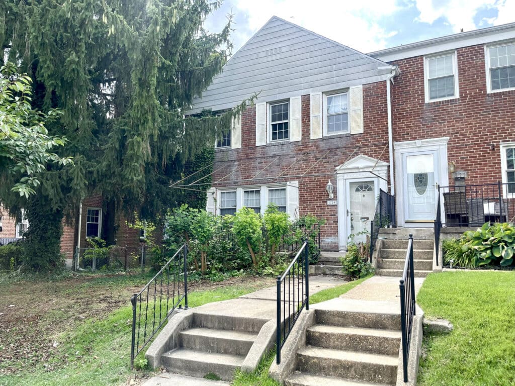 Front exterior of a two-story brick end-of-group townhome in Loch Raven Village with concrete steps, iron railings, and a shaded front yard.