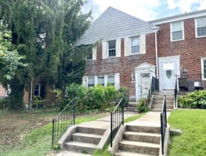 Front exterior of a two-story brick end-of-group townhome in Loch Raven Village with concrete steps, iron railings, and a shaded front yard.