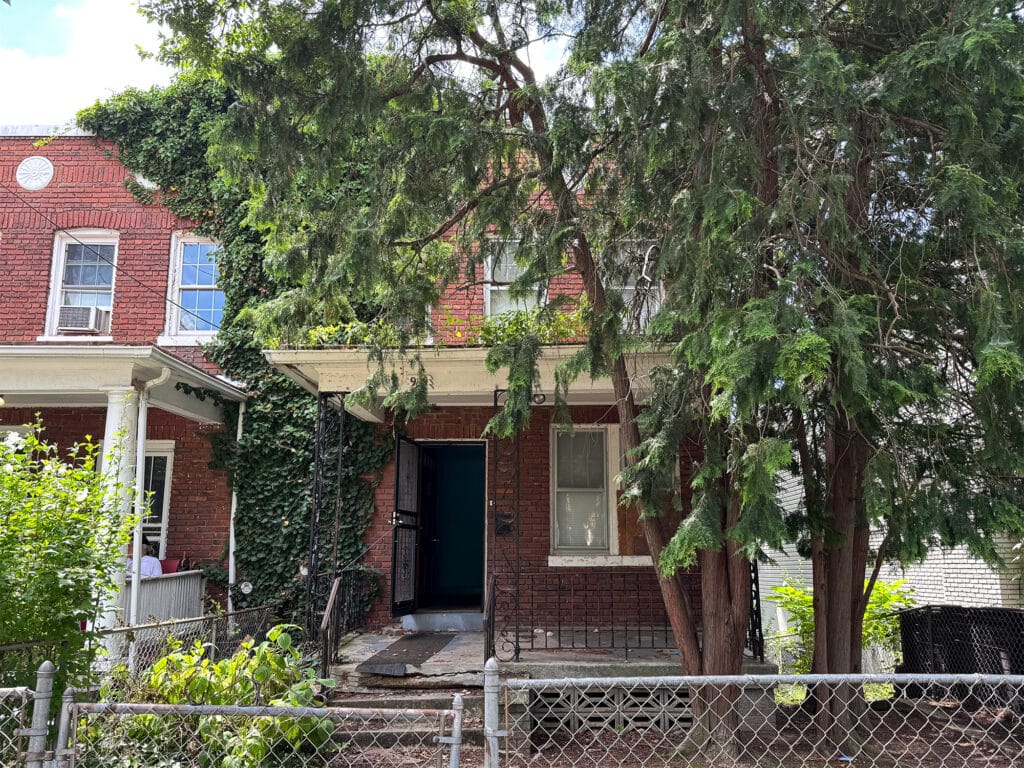 Front exterior of a semi-detached brick townhome at 909 Jackson Street NE in the Brookland neighborhood of Washington, DC, with a fenced yard and mature tree coverage.