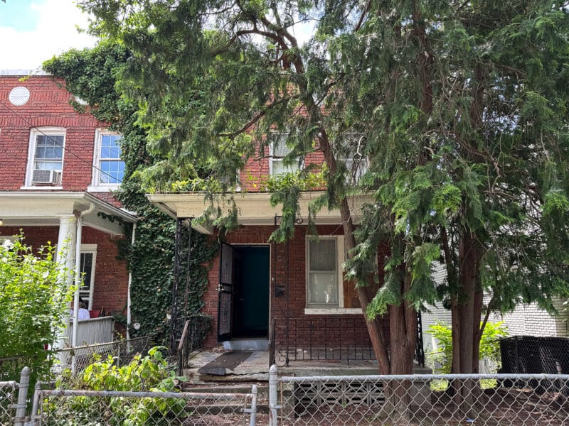 Front exterior of a semi-detached brick townhome at 909 Jackson Street NE in the Brookland neighborhood of Washington, DC, with a fenced yard and mature tree coverage.