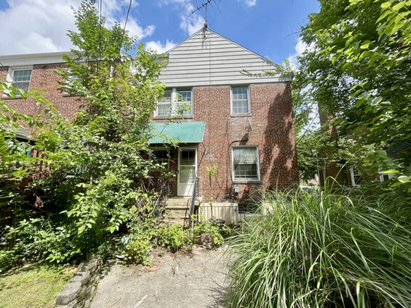 Rear exterior of a two-story brick townhome surrounded by overgrown vegetation, concrete patio, and shrubbery.