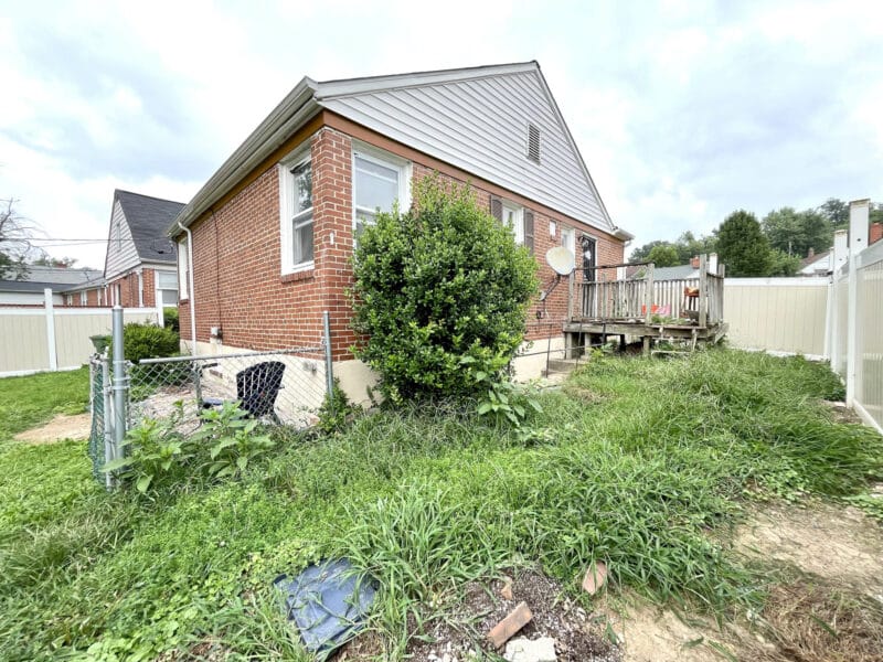 Side and rear view of 4902 Gilray Drive showing overgrown yard, chain-link fence, and rear deck.
