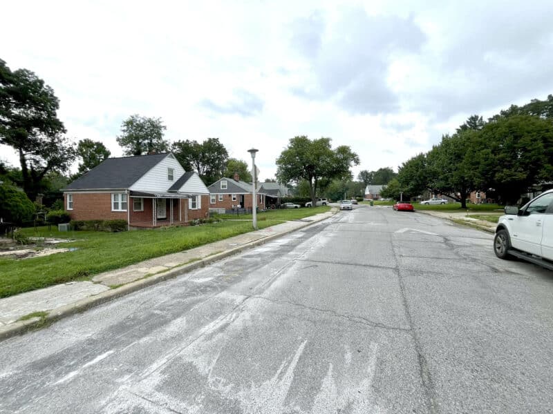 View of a residential street lined with one-story brick homes and grassy lawns in Northeast Baltimore.