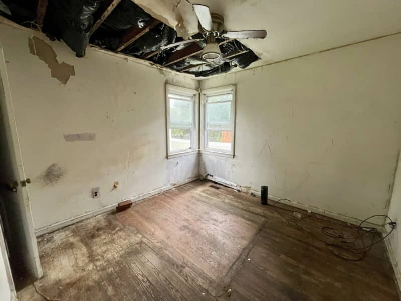 Damaged bedroom with exposed ceiling joists, white walls, and two corner windows at 4902 Gilray Drive.