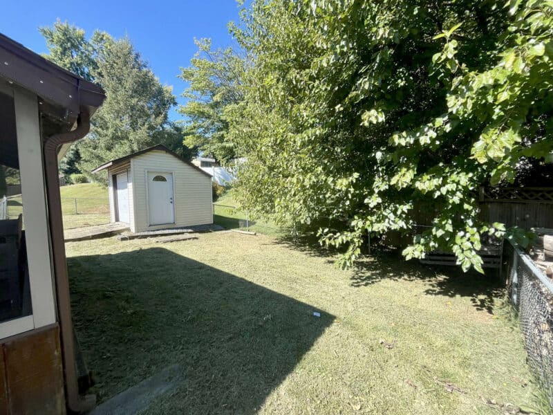 Backyard view featuring a storage shed, mature trees, and open grassy space enclosed by fencing.