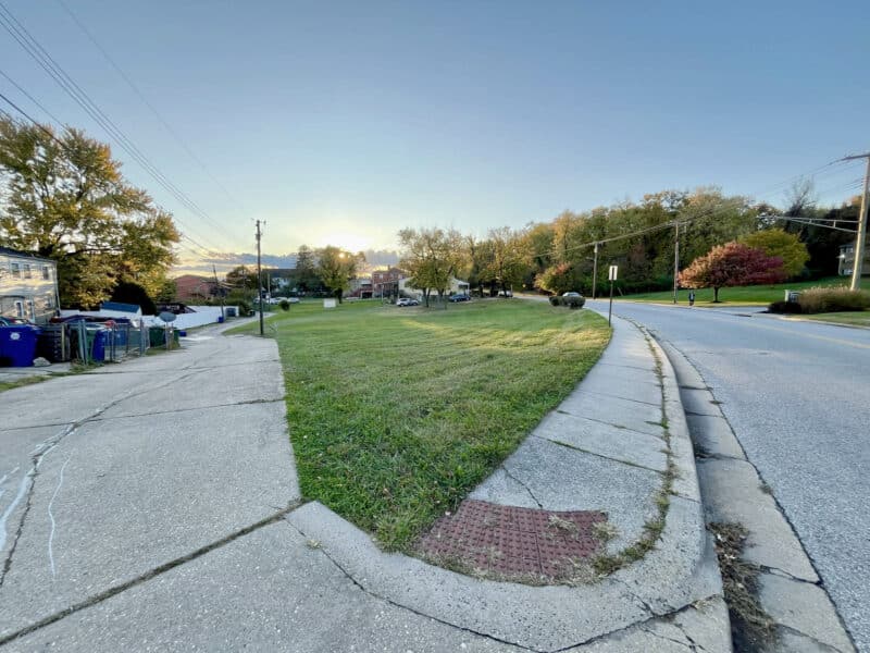 Wide view of the triangular 0.32-acre lot at 4611 Bowleys Lane, Baltimore, showing grass, sidewalk, and surrounding residential streetscape.