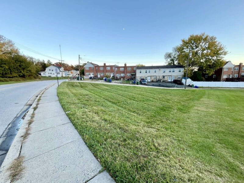View from the sidewalk looking across the grassed lot toward the rear of nearby rowhomes along Bowleys Lane.
