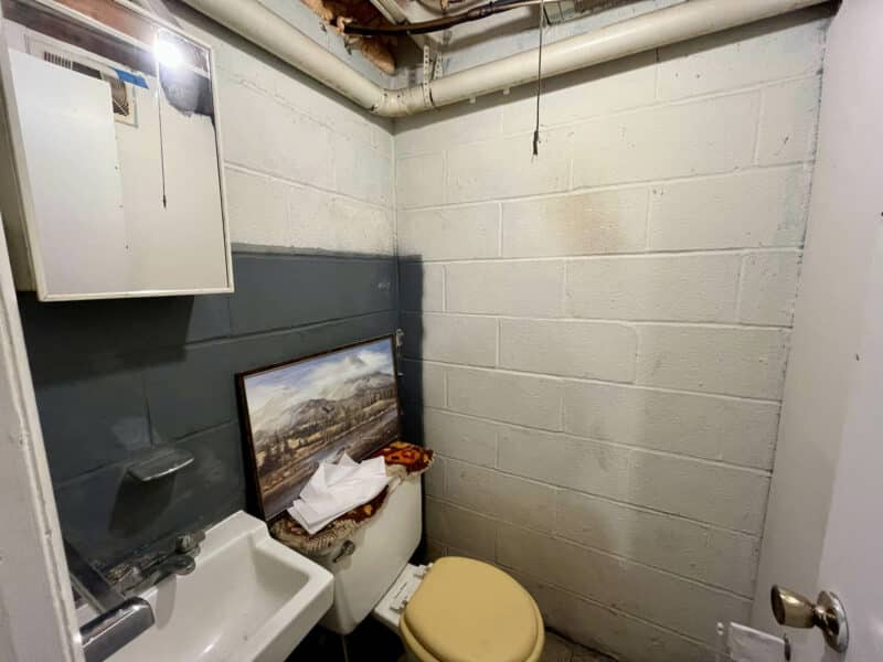 Basement half bathroom with exposed block walls, sink, toilet, and overhead plumbing.