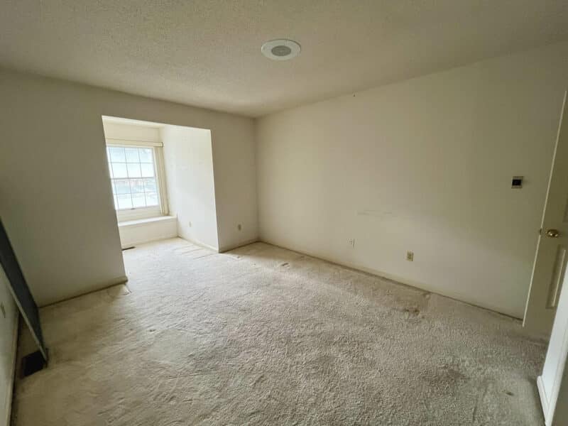 Bedroom with light carpet, single side window, and neutral walls needing cosmetic updates.