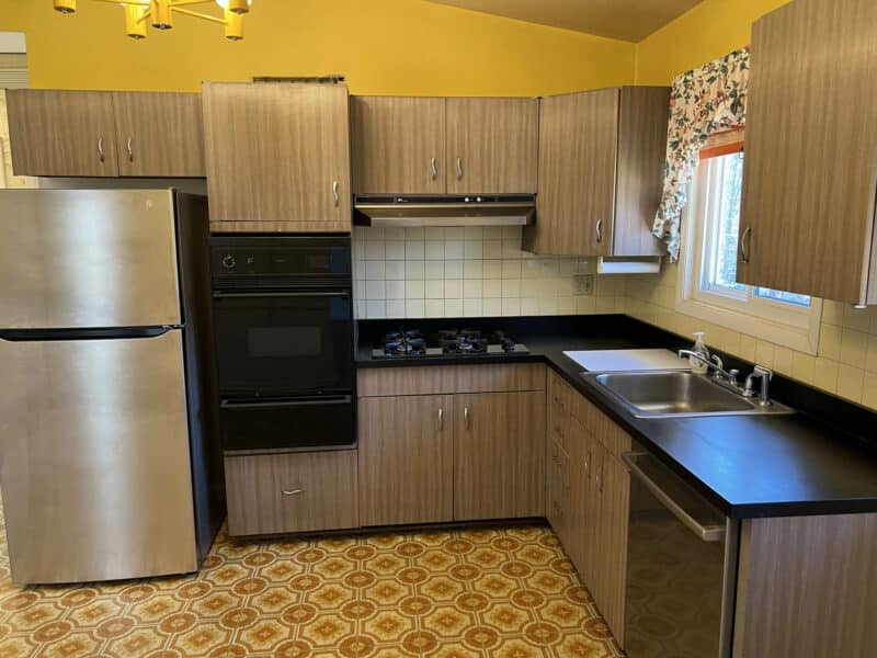 Mid-century style kitchen with wood-grain cabinets, stainless steel appliances, yellow tile backsplash, and retro patterned flooring.