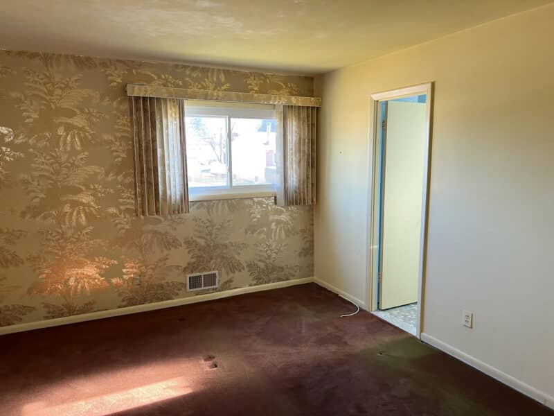 Bedroom with vintage patterned wallpaper, burgundy carpet, window with drapes, and doorway to an adjoining space.