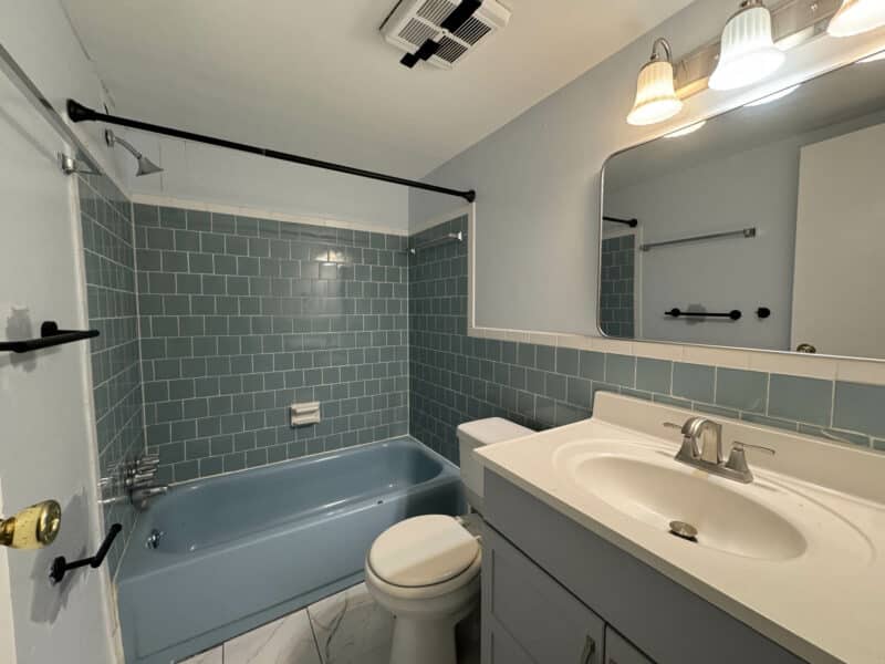 Bathroom with blue tile tub surround, white vanity, updated lighting, and ceramic tile flooring.