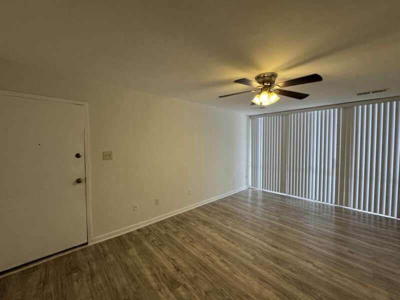 Alternate angle of the renovated living area showing vinyl plank flooring, entry door, and ceiling fan with natural light from vertical blinds.