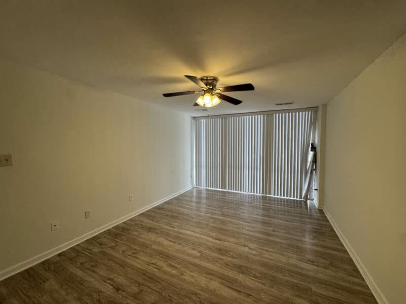 Wide-angle view of the living room showing updated vinyl flooring, ceiling fan, entry door, and natural light from balcony doors.