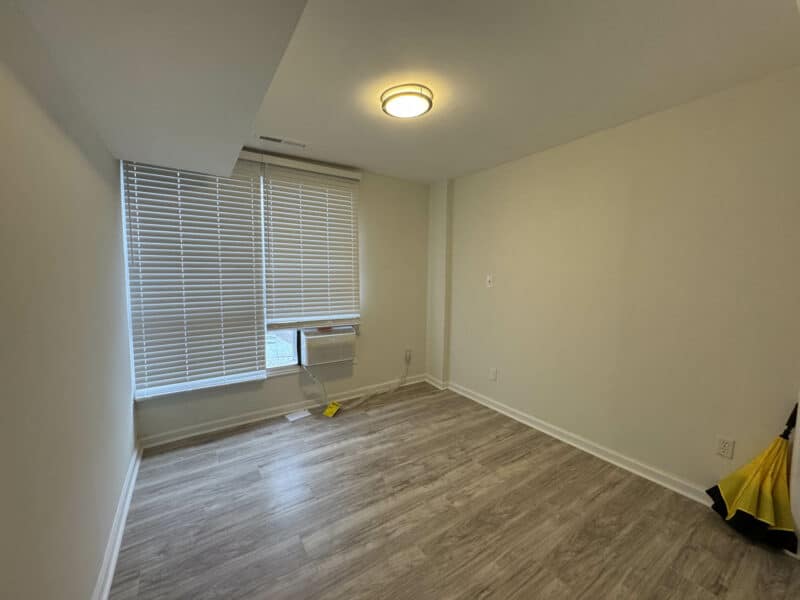 Bedroom with fresh paint, new vinyl flooring, window blinds, and window HVAC unit in the renovated Langley Park condominium.