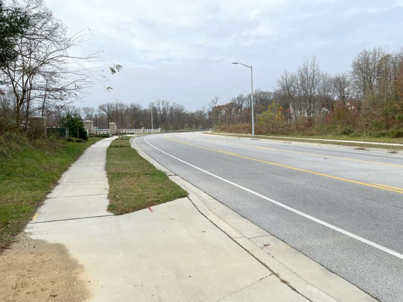 Street-level view of a curved road and sidewalk along Stokers Lane bordered by wooded areas on both sides.
