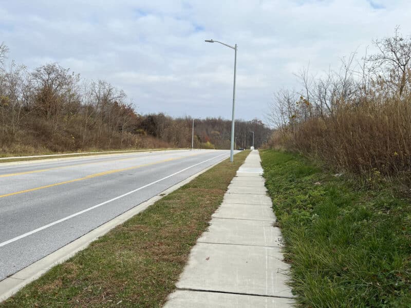 Straight sidewalk and roadway running alongside the wooded edge of the Stokers Lane parcel, with streetlights and open sky.