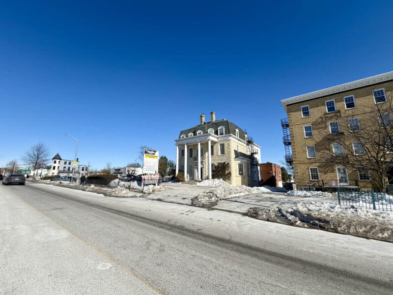 Wide-angle approach view of historic stone commercial building with columned entrance and on-site parking in winter conditions.