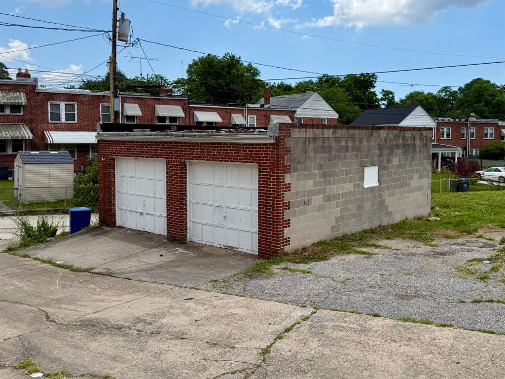 Two-bay concrete masonry garage building with white overhead doors and adjoining paved parking area in Baltimore's Belair-Edison neighborhood.