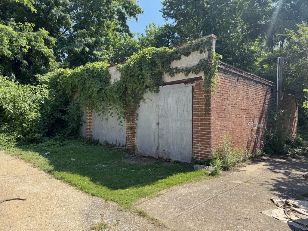 Corner exterior view of six-bay concrete masonry garage building with metal garage doors, mature trees, and dual alley access in Baltimore's Walbrook neighborhood.
