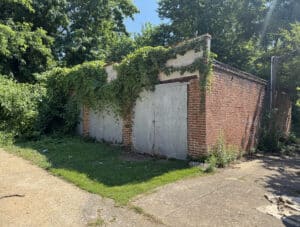 Corner exterior view of six-bay concrete masonry garage building with metal garage doors, mature trees, and dual alley access in Baltimore's Walbrook neighborhood.
