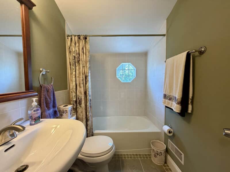 Upper-level full bathroom with tiled tub and shower surround, pedestal sink and octagonal window.